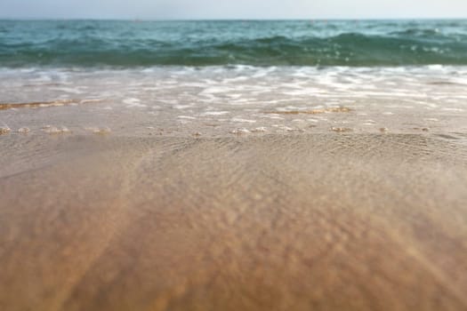Low angle photo from ground level - beach sand wet from sea, drops of water in air, small waves and blurred ocean in distance. Abstract vacation background.