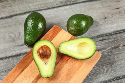 Tabletop view, chopping board on gray wood desk, avocado cut in half, with seed visible, and two whole pears next to it.