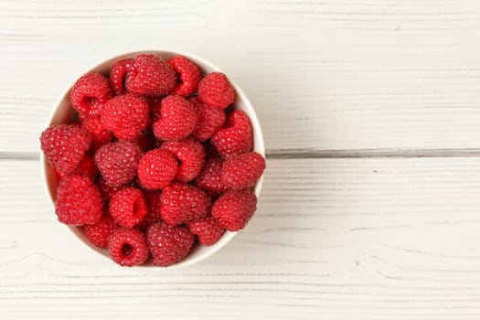 Tabletop view, small porcelain cup filled with raspberries on white boards desk.