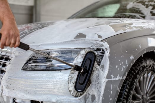 Front light of silver car washed in self serve carwash, man hand holding brush,  cleaning the glass with shampoo