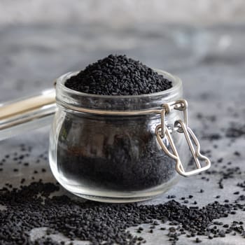 Indian spice Black cumin (nigella sativa or kalonji) seeds glass jar on grey table close up. Traditional asian medicine, healthy and vegetarian food concept