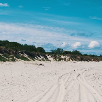 Dunes, overgrown with grass in places, car tracks on the sand. Blue sky with clouds.