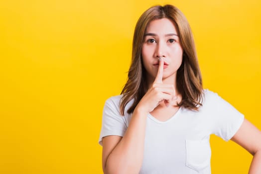 Asian Thai happy portrait beautiful cute young woman standing wear white t-shirt making finger on lips silent quiet gesture looking to side, studio shot isolated on yellow background with copy space