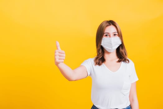 Portrait Asian Thai beautiful happy young woman wearing mask protection, show thumb finger good stopping virus outbreak control health care, studio shot isolated on yellow background, with copy space
