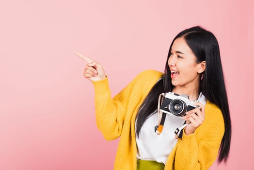 Attractive energetic happy Asian portrait beautiful cute young woman teen excited smiling holding vintage photo camera and pointing finger to side space, studio shot isolated on pink background