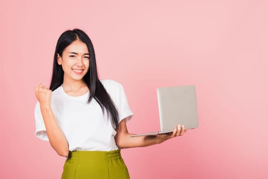 Portrait of happy Asian beautiful young woman teen confident smiling face holding using laptop computer and excited celebrating success, studio shot isolated on pink background, with copy space