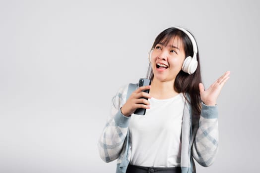Woman excited smiling listening to music radio in bluetooth headphones and singing song on smartphone studio shot isolated white background, happy Asian young female Karaoke online app in mobile phone