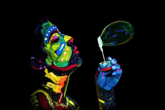 Girl painted with UV colors blows a soap bubble against black background