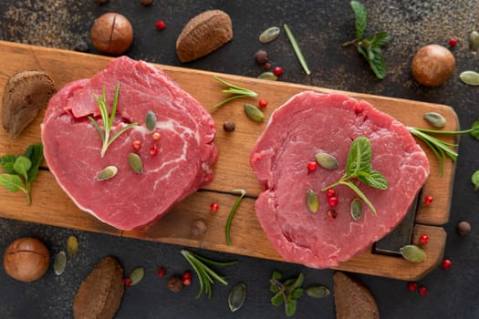 Raw beef fillet steaks on wooden board on dark table top view. Portioned filet mignons with spices and herbs. Concept of summer food