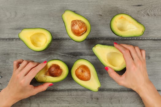 Tabletop view, woman hands with red nails holding avocados cut in half, more halves, some of them with seed around on gray wood desk.
