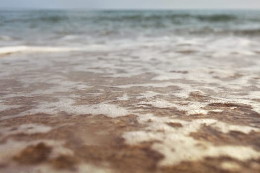 Low angle photo from ground level - beach with shallow water and white foam over sand, wet drops in air, blurred ocean in distance. Abstract vacation background.
