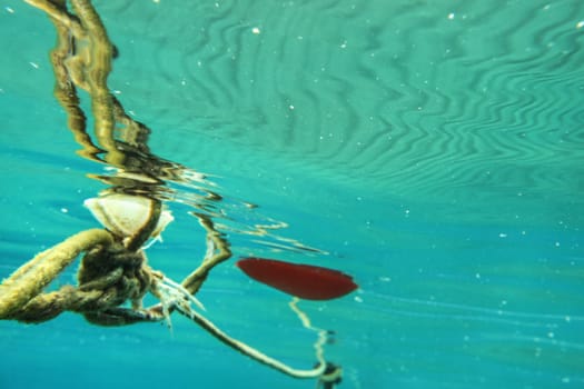 Underwater photo, old buoy and rope, distorted by water refraction, waves on surface creates wavy pattern. Abstract marine background with space for text on the right.