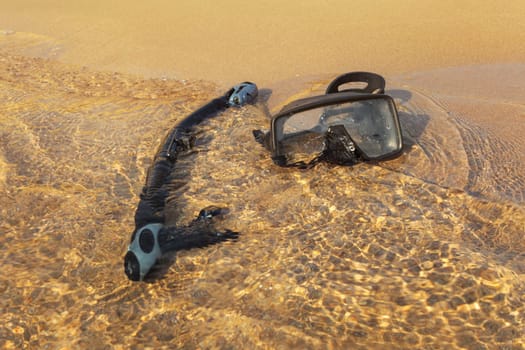 Black snorkel breathing tube and diving mask, in shallow sea on fine sand beach, sun shining on clear water.