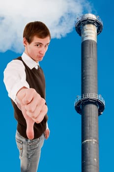 Waist up view of a young white male showing thumbs down next to a smoking factory chimney against a blue sky.