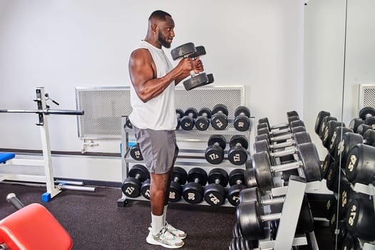 An African American man shakes his biceps with dumbbells while standing in front of a mirror. High quality photo