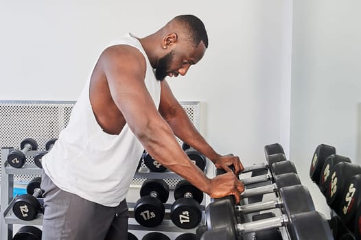 An African American man stands in front of a mirror and leans on dumbbells in the gym. High quality photo