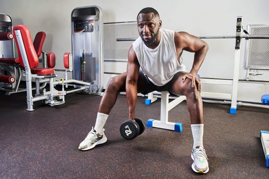An African American man shakes his biceps while sitting on a bench in the gym. High quality photo