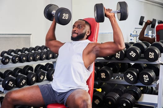 An African-American man does a dumbbell press while sitting on a bench in the gym. High quality photo