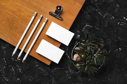 Branding identity mockup. Blank business cards, pencils, succulent plant, metal clip and wooden board on black plaster background. Flat lay.