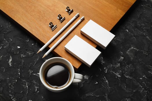 Coffee cup and blank stationery: business cards, pencils and metal clips on wooden board background.