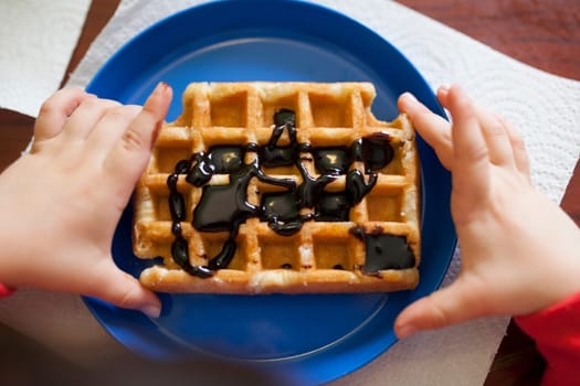 Child holds a waffle over blue plate. Waffled is filled with chocolate syrup