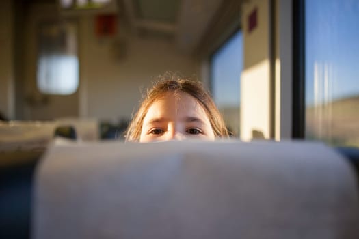Child girl peeking out from behind a headrest train seat. Inside the train carriage