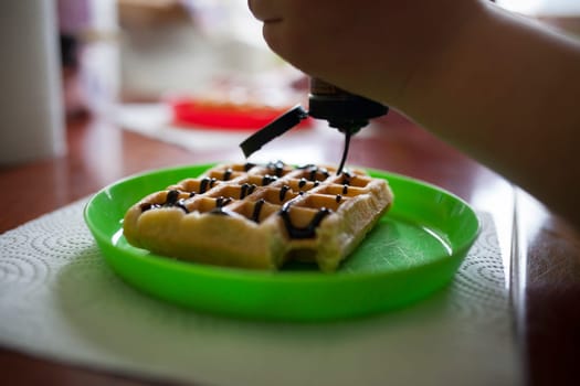 Child pouring chocolate syrup on a waffle. Closeup