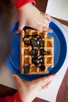 Child holds a waffle over blue plate. Waffled is filled with chocolate syrup