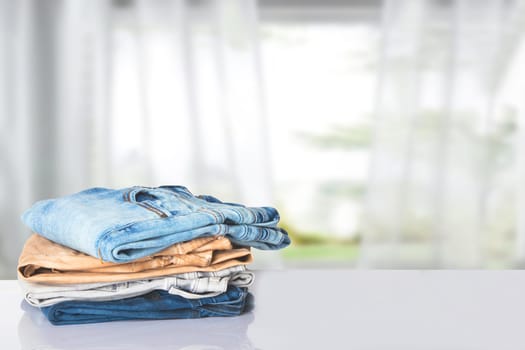 Stack of jeans. Closeup of a pile of colorful female denim pants on a bright table against abstract blurred curtain background. Copy space.