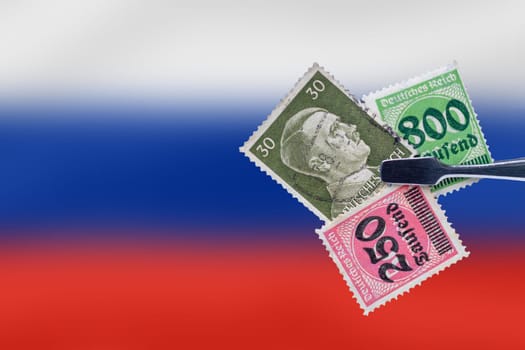 A pair of tweezers holds old stamps of the Third Reich, one stamp with the image of Hitler and two other re-stamped inflation stamps over abstract blurred white, blue and red background. Space.