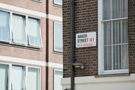 Baker Street sign affixed on dark brick wall next to drainage pipe and empty window. There are other building windows in the blurred background.