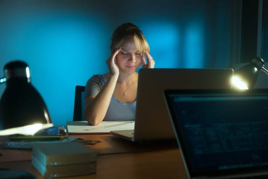 Beautiful woman working as interior designer, staying late at night in office with drawings and laptop computer. The girl feels tired and her eyes hurt.