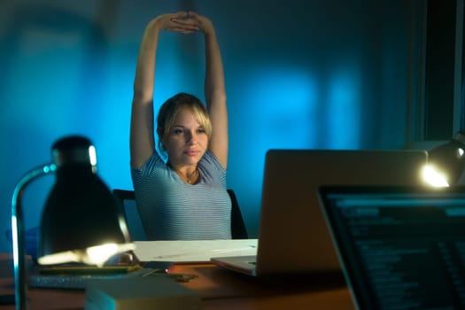 Beautiful woman working as interior designer, staying late at night in office with drawings and laptop computer to complete a project. The girl yawns and stretch her arms