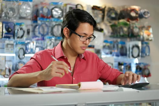 Young asian entrepreneur owning a computer shop and checking bills and invoice with calculator, doing budget and reviewing expenses.