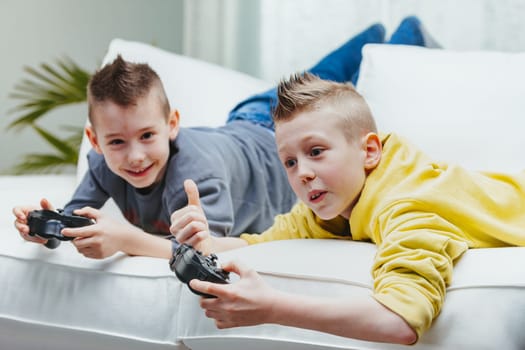 Two young brothers playing video games together on the couch with one giving a thumbs up gesture of success watched by his grinning sibling