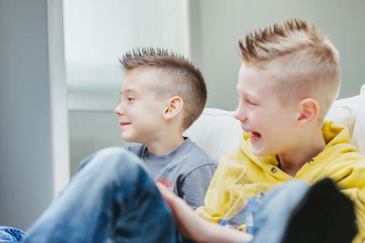 Cute young boy with modern spiky hairstyle sitting on a sofa with his laughing brother as they watch or play games at home