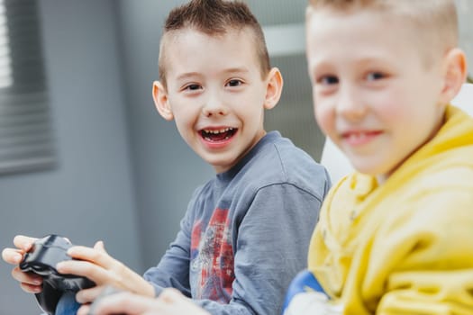 Young boy playing video games on the sofa alongside his brother or friend turning to laugh at the camera in a candid portrait