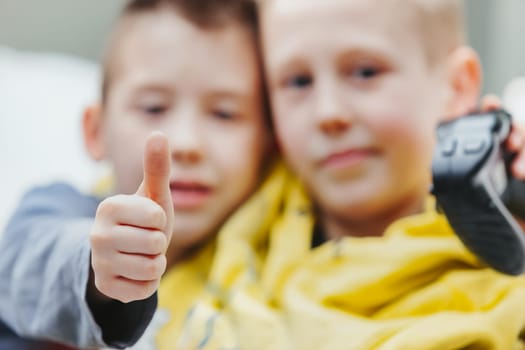 Young boy giving a thumbs up gesture of success as he sits close together with his brother or friend playing video games with focus to his hand