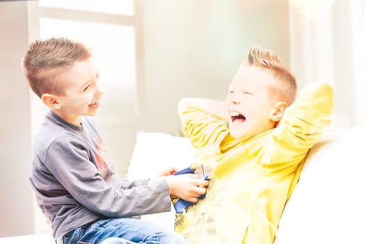 Two laughing young brothers tickling each other on a sofa as they relax having fun in a high key portrait