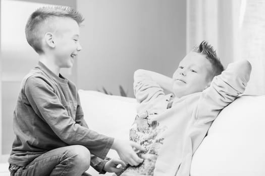 Greyscale image of two young brothers having fun playing together on a sofa at hole laughing and tickling each other