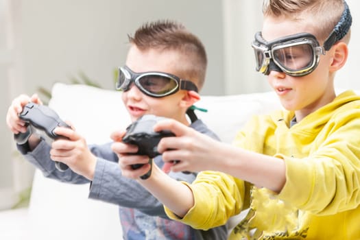 Two competitive young boys playing computer games seated side by side on a sofa with raised games controllers wearing goggles