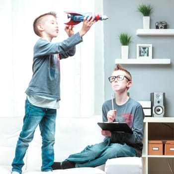 Active young boy playing with his toy spacecraft indoors at home watched by his older brother with a pensive expression