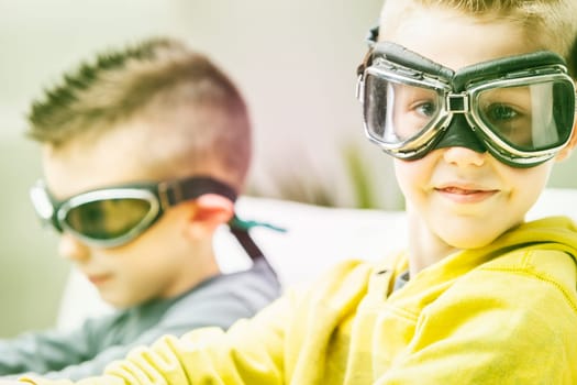 Cute high key portrait of a young boy wearing goggles turning to grin at the camera as he relaxes with his brother or friend on a sofa