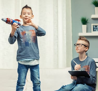 Two cute young boys with Mohican spiky hairstyles playing in a living room with a toy rocket with focus to a youngster standing grinning at the camera