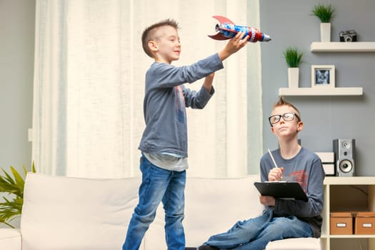 Young boy playing with a colorful toy rocket standing on the sofa in the living room watched by his older brother working on homework on a clipboard