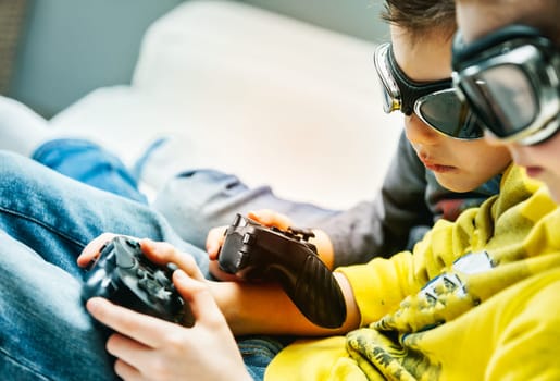 Young brothers wearing goggles huddling together playing video games on a sofa in a close up cropped view with focus to their hands and the controllers