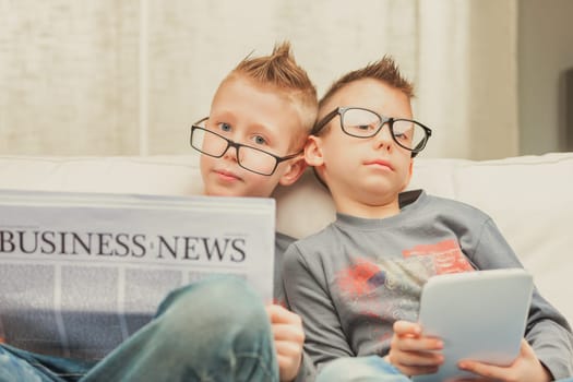 Two studious young brothers wearing glasses relaxing on a sofa, one with the Business Times and one with a tablet computer