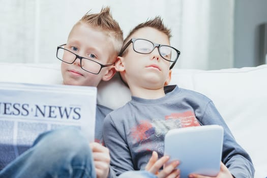 Portrait of two cute young boys in glasses sat at home with tablet and business newspaper