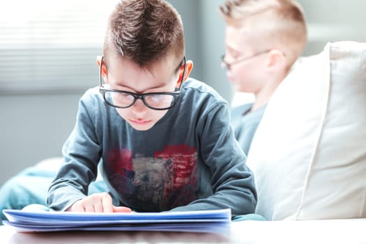 Studious young boy wearing glasses lying on a sofa reading a newspaper in a close up head to toe view in a lifestyle concept
