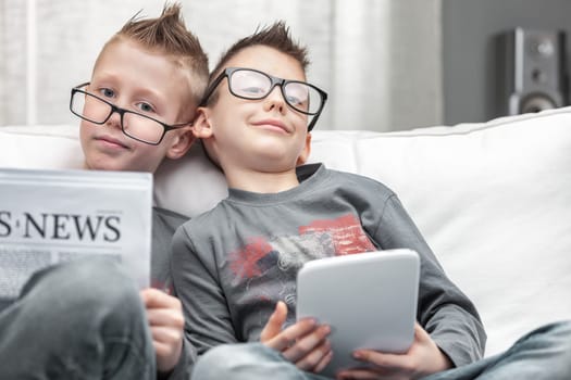 Two young boys wearing eyeglasses relaxing on a sofa together with tablet-pc and Business Times newspaper smiling happily at the camera